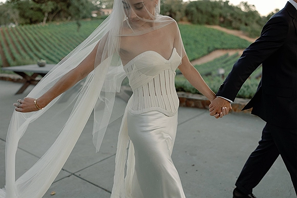 Wedding couple portrait of bride and groom holding hands, her long veil blowing behind a strapless corset gown on a vineyard path