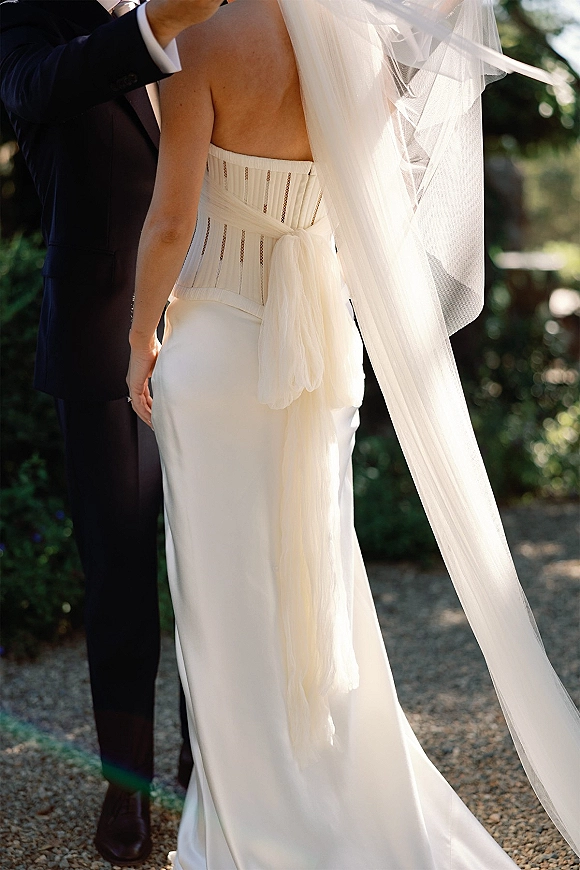 First look moment as groom lifts the wedding veil over a bride in a strapless corset dress on a sunlit gravel path amid garden greenery