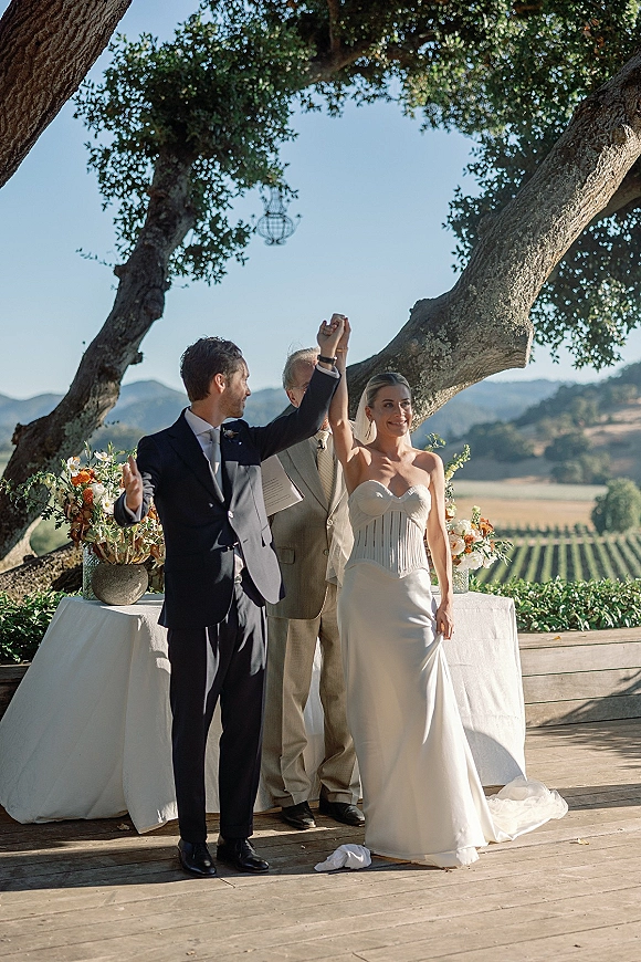 Recessional moment as newlyweds holding hands raise their arms on a wooden deck under a large oak, lantern above and vineyard hills behind