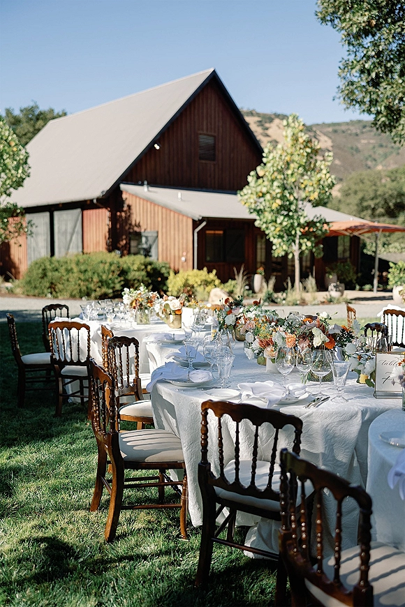 Outdoor reception tables set with white linens, wooden chairs, and floral centerpieces on a lawn with a barn and hills beyond