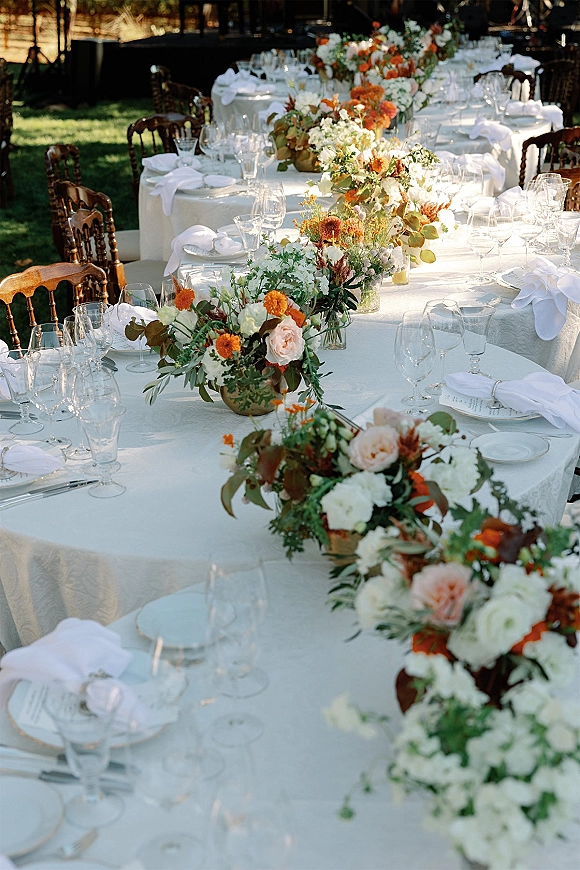 Reception tablescape with outdoor wedding reception tables dressed in white linens, floral centerpieces, greenery, and clear goblets on a lawn under trees