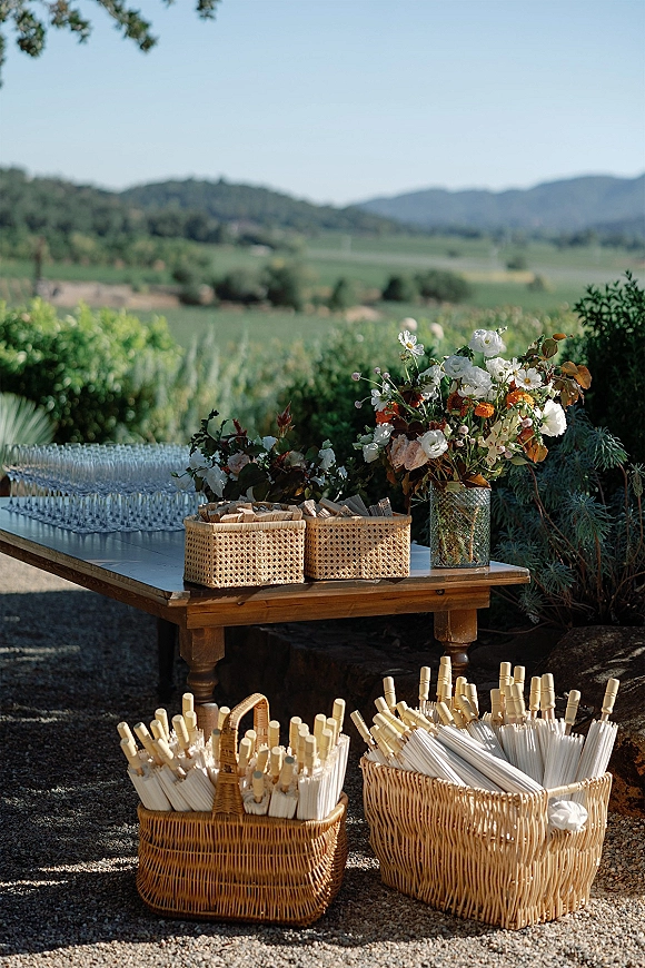 Wedding welcome table with wedding welcome table decor featuring glassware, wicker baskets of confetti wands, and florals in a vase in a vineyard setting