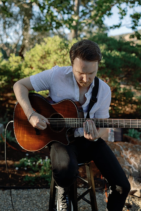 Wedding musician playing acoustic guitar with a slide, seated in a sunlit garden with trees, greenery, and gravel ground behind