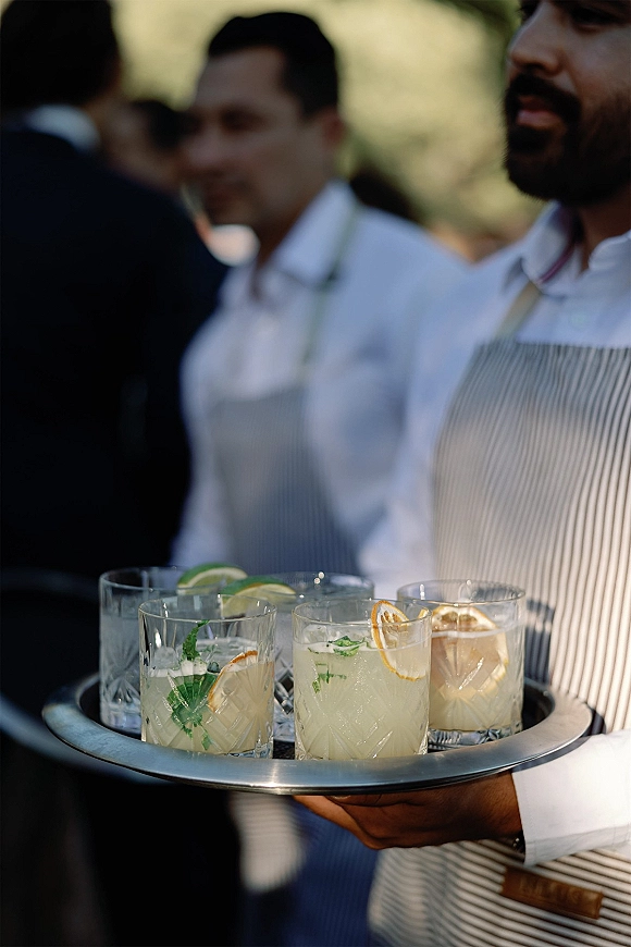 Wedding cocktail service with a server holding a metal tray of signature wedding cocktails in rocks glasses with citrus and mint, outdoors amid greenery