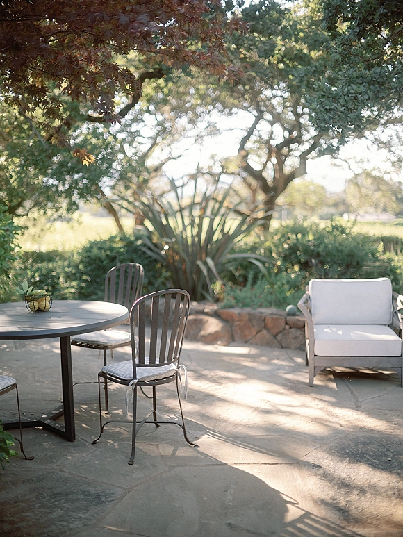 Outdoor patio seating with garden patio furniture around a round table and white sofa, fruit bowl centerpiece, sunlit trees and stone wall backdrop