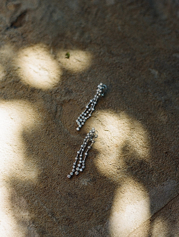 Bridal earrings with crystal drop earrings in metal settings resting on a stone surface, dappled sunlight casting soft shadows