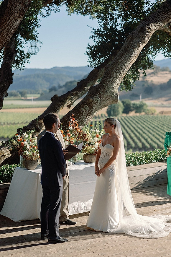 Wedding vows as bride in strapless dress and cathedral veil faces groom on a wooden deck under an oak tree with vineyard rows beyond