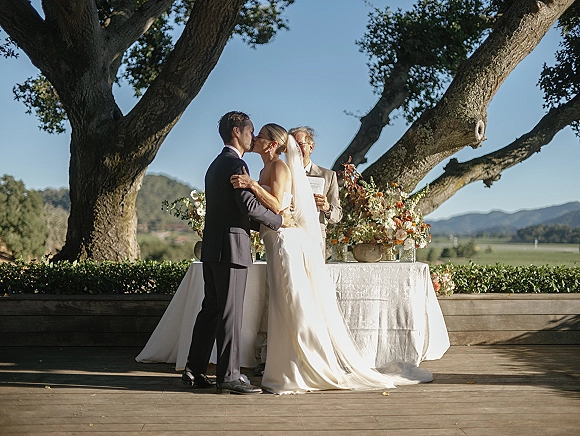 Wedding kiss during an outdoor wedding ceremony, bride in long veil and satin gown kissing groom by a flowered table under oak trees