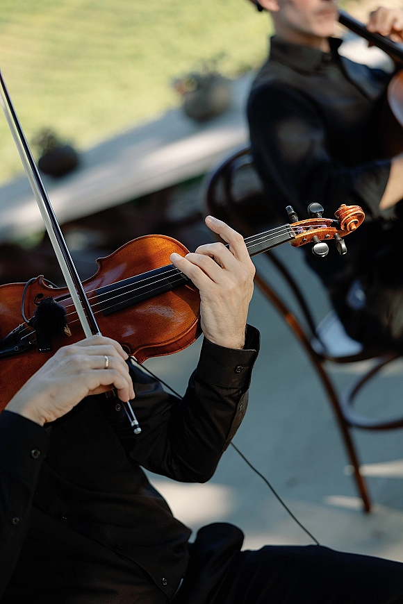 Wedding musicians play live violin music, bowing an amplified violin near a music stand on an outdoor patio beside the lawn