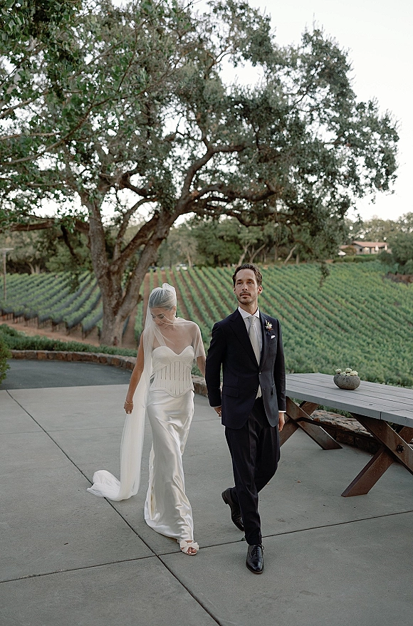 Couple portrait of bride and groom walking hand in hand, her long veil flowing beside vineyard rows under a large oak tree