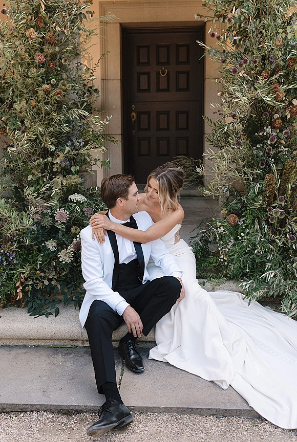 Couple portrait of bride hugging groom on stone steps, her strapless long-train dress beside his white dinner jacket under greenery florals
