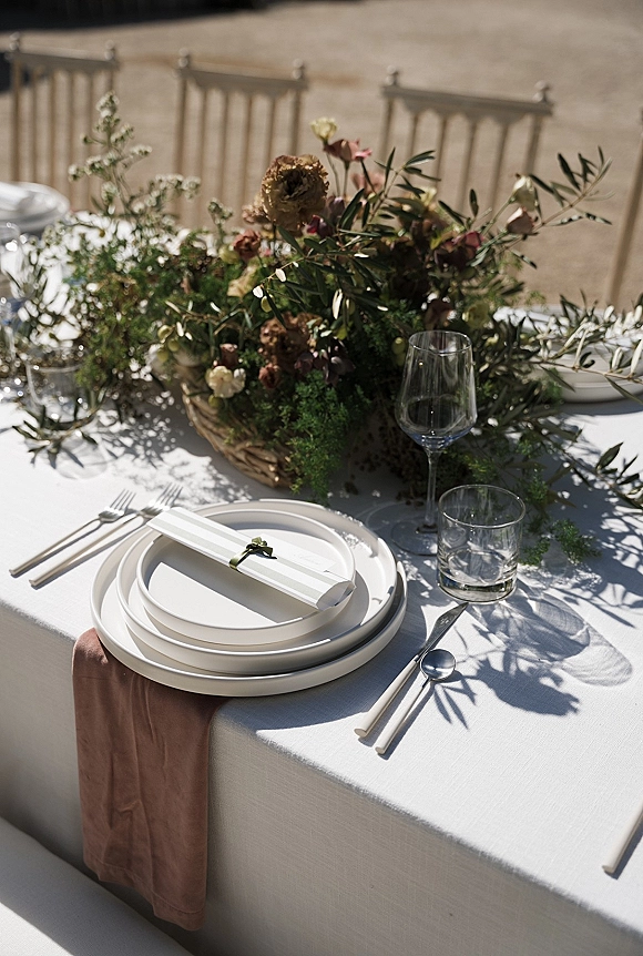 Reception tablescape with wedding place setting on white linens, brown runner, ribbon-tied menu, ceramic plates, and greenery centerpiece outdoors
