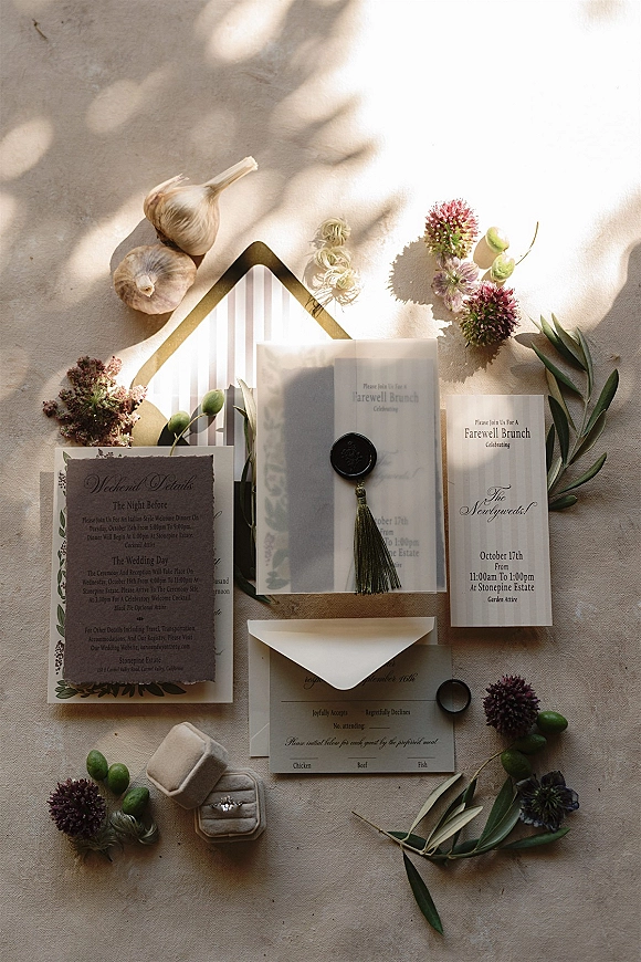 Invitation suite wedding invitation flat lay with wax seal, tassel, RSVP and details cards on linen in dappled light, with olive branches and purple flowers