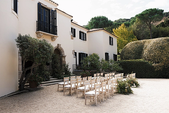 Ceremony setup with wood chairs and white cushions facing a villa’s arched doorway, with greenery and low aisle florals in a gravel courtyard