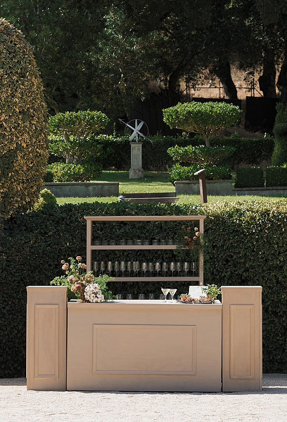 Wedding bar setup with outdoor wedding bar shelves, champagne flutes, cocktail glasses, citrus garnishes, and floral accents in a garden setting