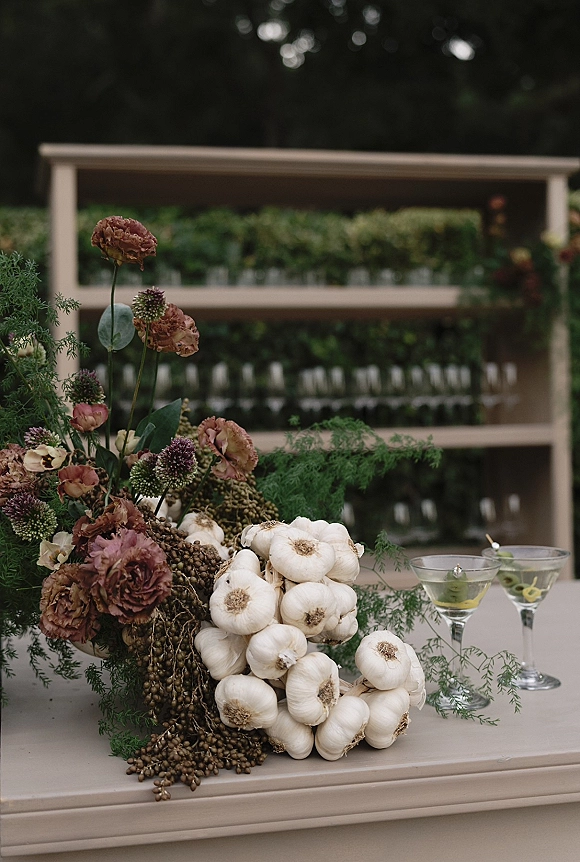 Wedding bar decor with a floral arrangement and greenery on the bar top beside cocktail glasses with olive garnish, bar shelves behind