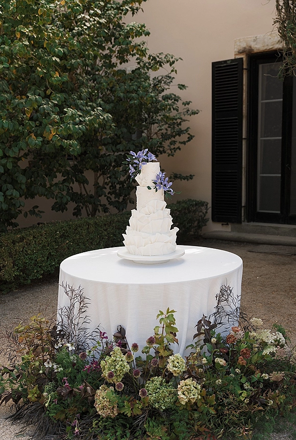 Wedding cake with textured buttercream tiers and a purple flower topper on a pedestal table, set on a gravel patio by a stucco wall