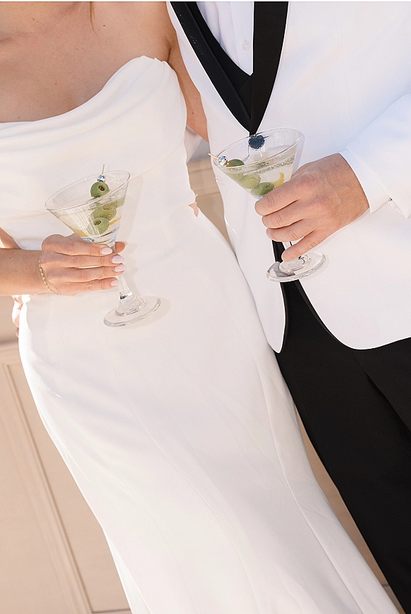 Wedding cocktails held by bride and groom, martini glasses with olive garnish and picks, showing bridal gown, white tuxedo jacket, black lapel