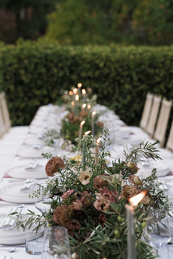 Reception tablescape on a long banquet table with floral garland, taper candles, white linens, place cards, and hedge backdrop
