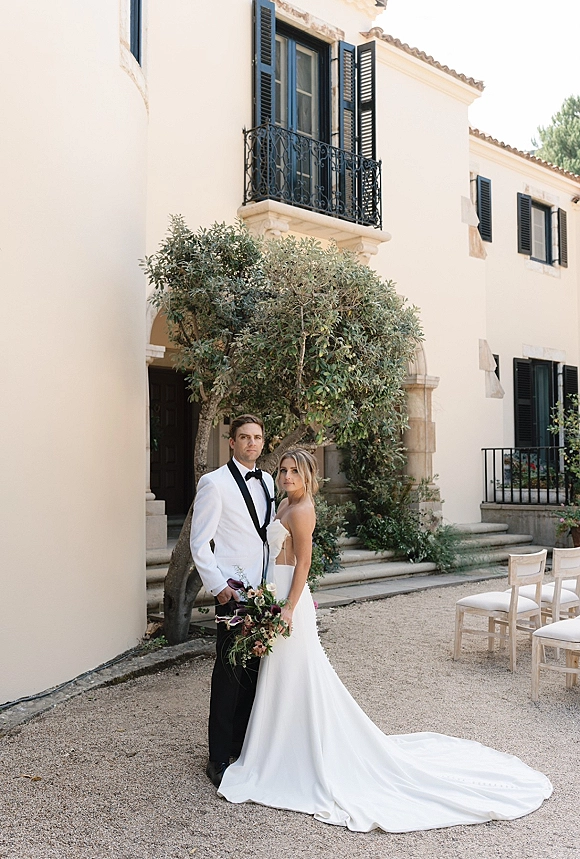 Couple portrait of bride holding wedding bouquet with greenery, standing on stone steps by a villa facade with black shutters and balcony