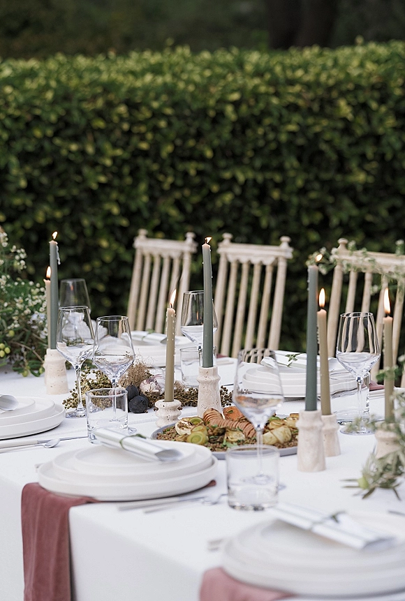 Reception tablescape with taper candles in stone holders, white plates, folded napkins, and glassware on a mauve runner beside garden hedges