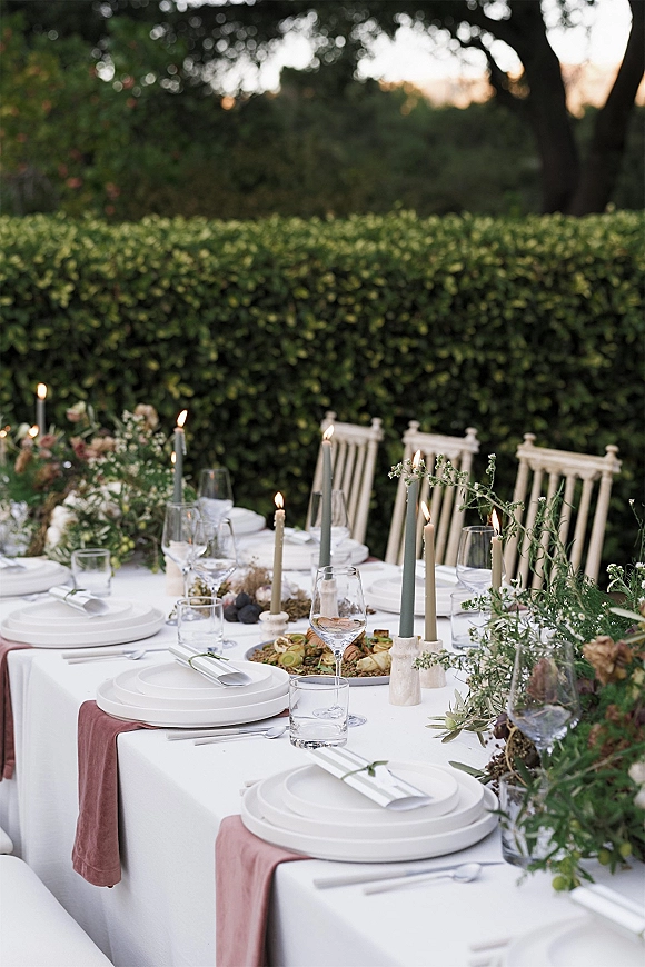 Reception tablescape with white tablecloth and blush runner, taper candles and floral garland on a long banquet table at sunset in a garden hedge setting