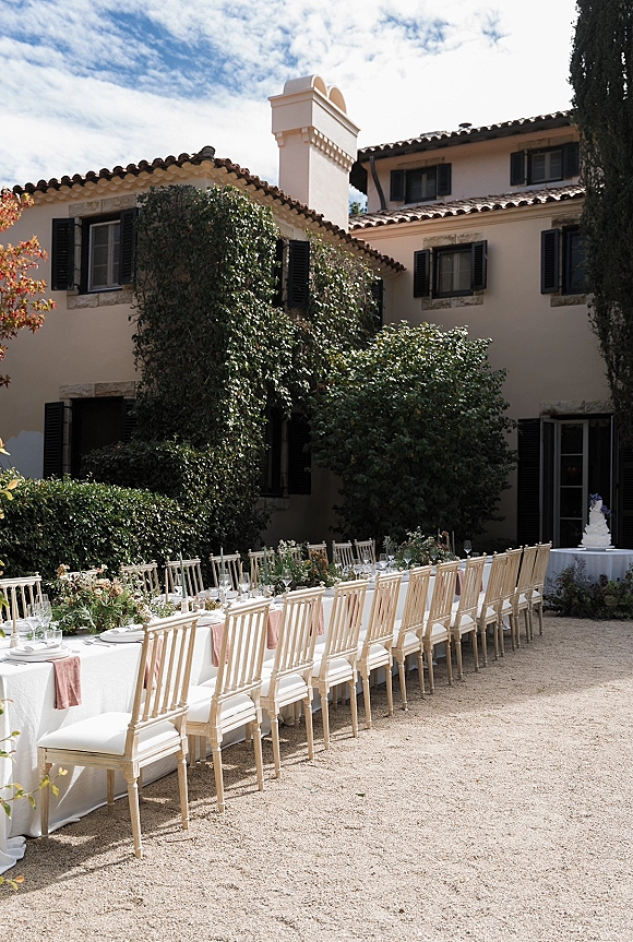Outdoor reception tablescape with a long banquet table wedding setup, pink napkins and greenery garland in a villa gravel courtyard