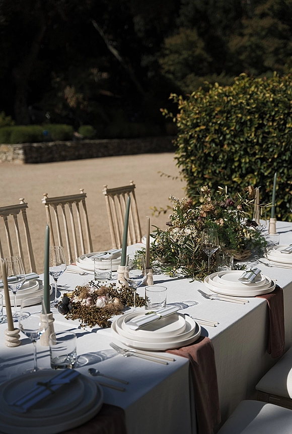 Reception tablescape with white tablecloth, taper candles and low floral greenery centerpiece on a long banquet table on a gravel patio