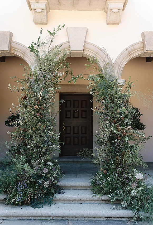 Wedding entrance decor with lush greenery and floral doorway installation framing stone steps, branches spilling around an arched double door