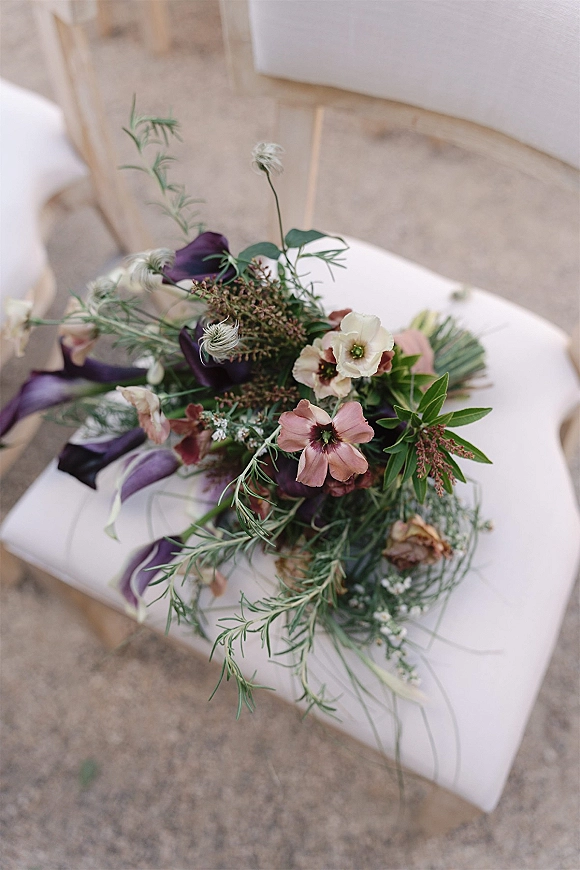 Wedding bouquet with purple calla lilies, blush blooms, and greenery resting on a white chair, with blurred outdoor ceremony seating behind