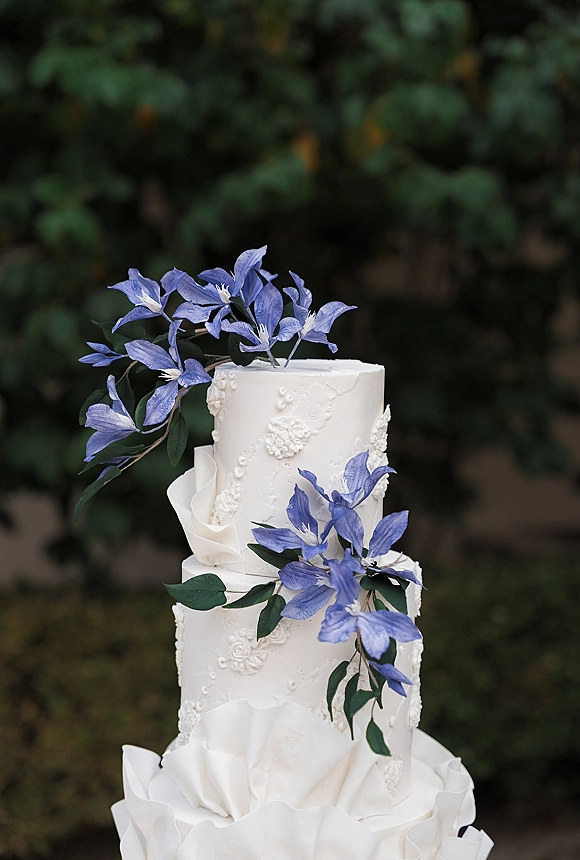Wedding cake with textured white tiers and cascading blue sugar flowers and green leaves, set against softly blurred outdoor greenery
