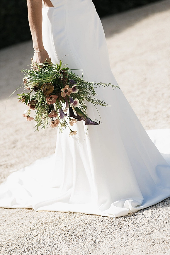 Bridal bouquet wildflower wedding bouquet with pink blooms and purple calla lilies, ribbon-wrapped stems in hand over sunlit pavement
