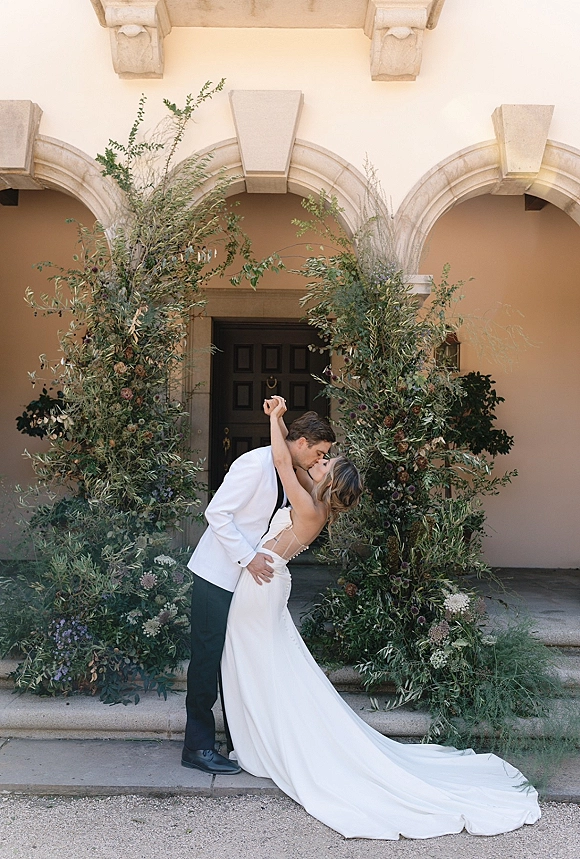 Wedding kiss portrait of bride and groom kissing in a dipped pose, her long train flowing by a greenery arch on stone steps