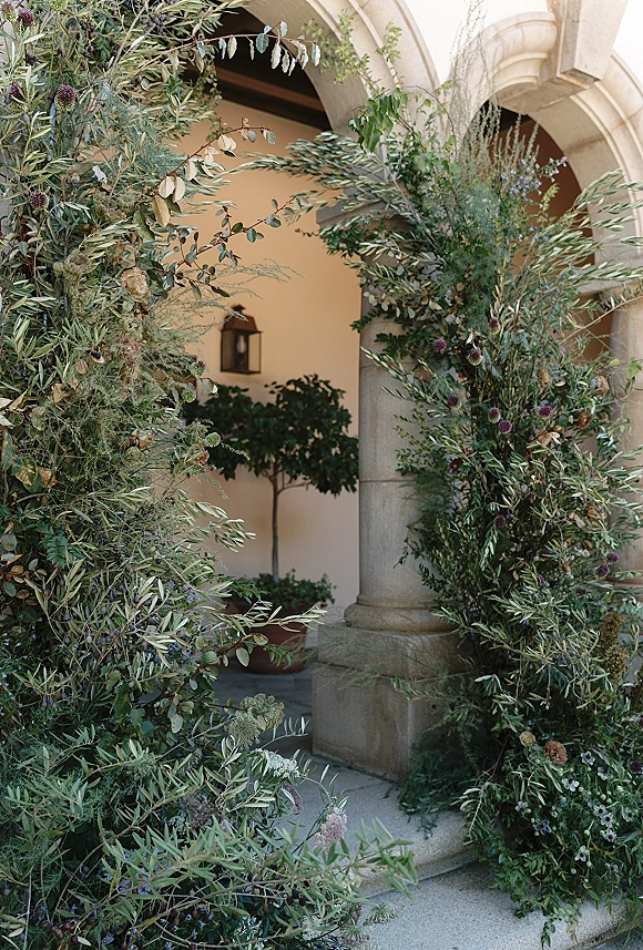 Floral ceremony arch with greenery wedding arch design, thistle and eucalyptus garland framing a stone archway with hanging lantern in courtyard