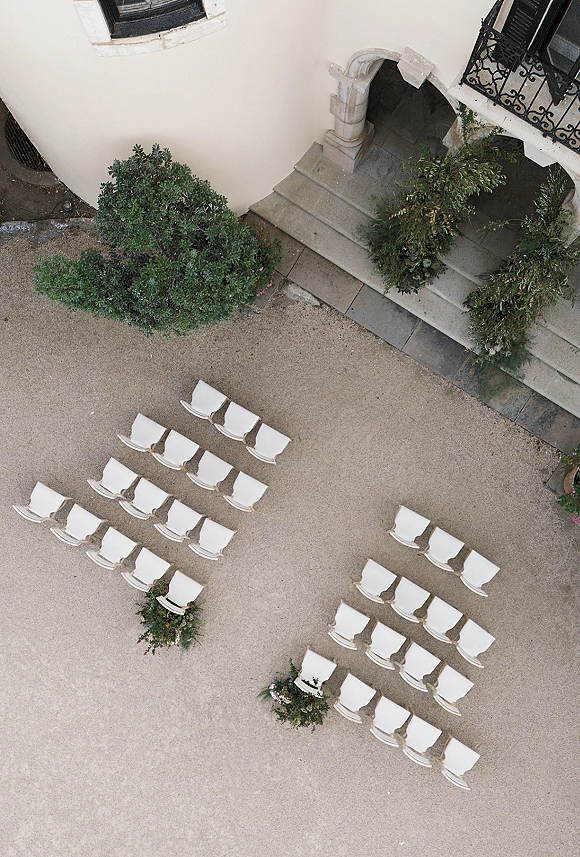 Outdoor ceremony setup with ceremony seating layout of white chairs and greenery florals in a gravel courtyard by stone steps and archway