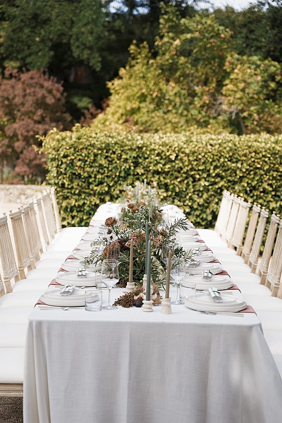 Reception tablescape with a long banquet table setup, white linens, silver flatware, taper candles, and greenery garland by a hedge outdoors