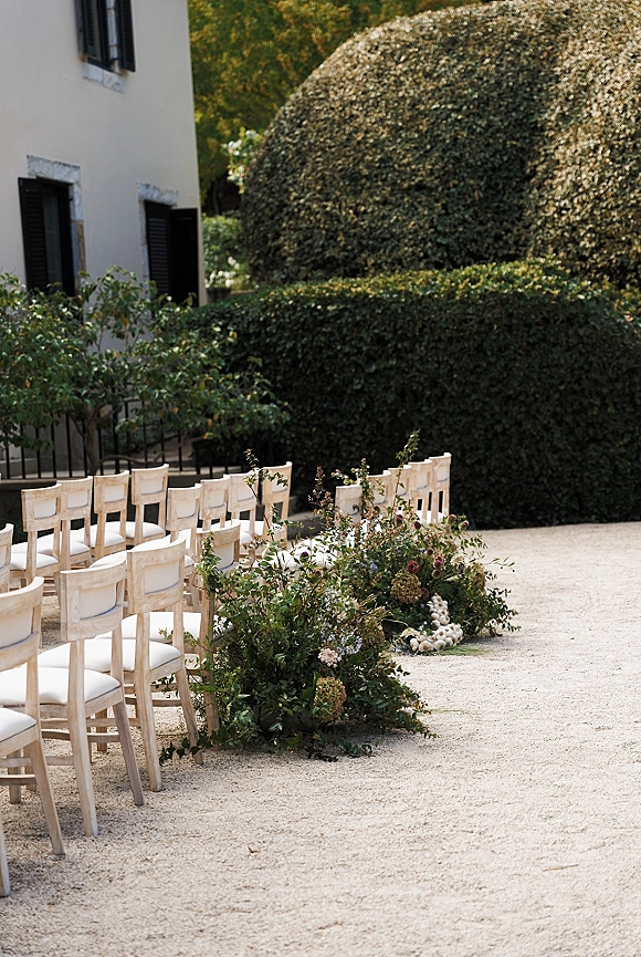Outdoor ceremony setup with garden ceremony seating, wooden chairs and hydrangea aisle florals in a gravel courtyard by a white stucco building
