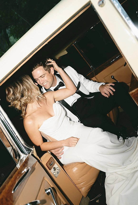 Couple portrait of bride and groom in car, bride in strapless gown leaning on smiling groom in white tux, tan leather interior behind