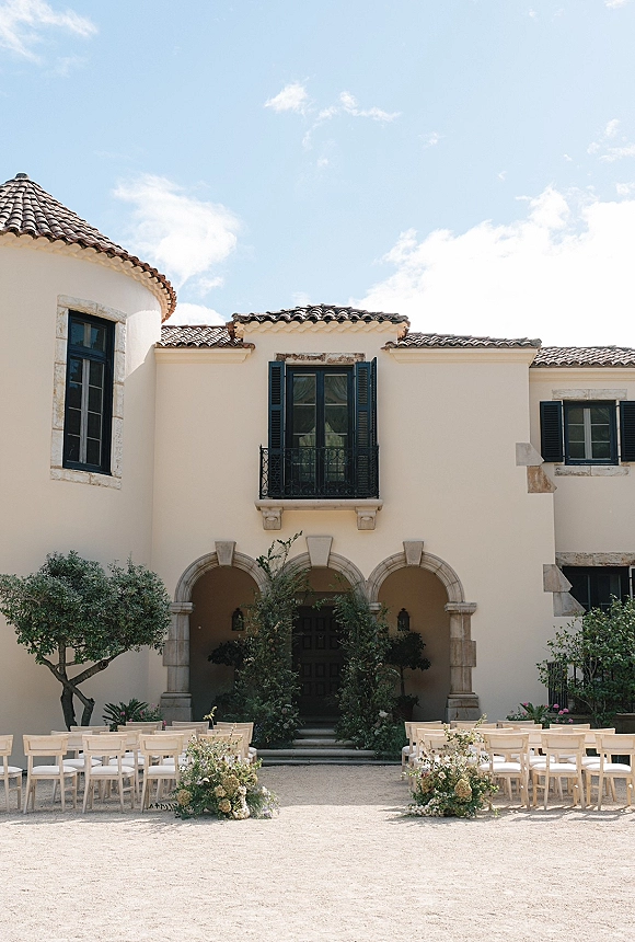 Ceremony setup with wood ceremony chairs flanking a floral-lined aisle in a villa courtyard, framed by stucco arches and terracotta roof tiles