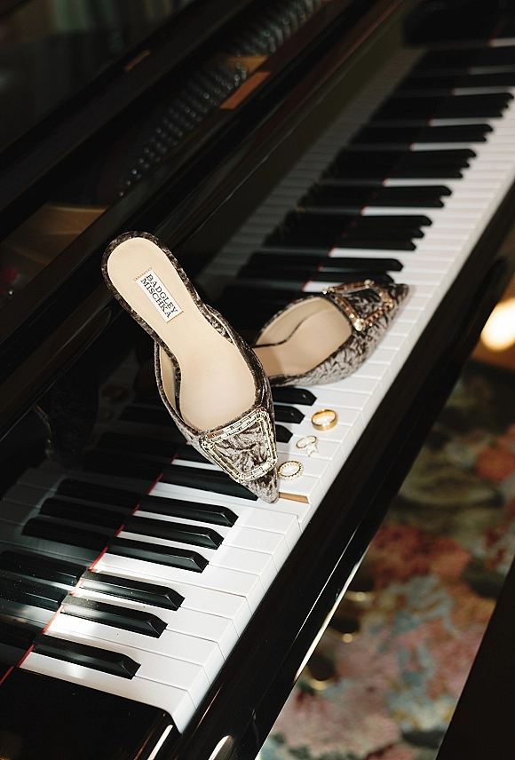 Bridal shoes with jeweled buckles in a wedding details flat lay on grand piano keys with rings and earrings on a patterned rug