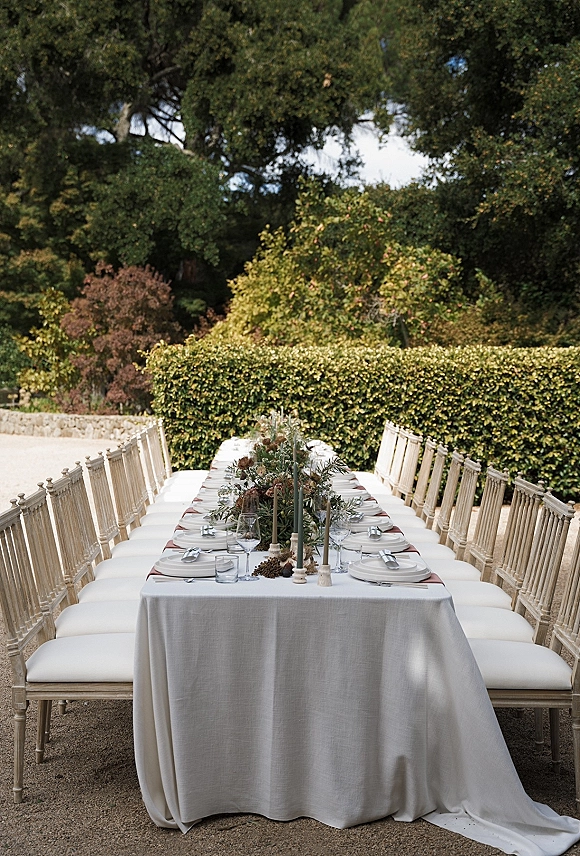 Reception tablescape with a long banquet table wedding setup of white linen, wooden chairs, greenery garland, and taper candles in a garden setting
