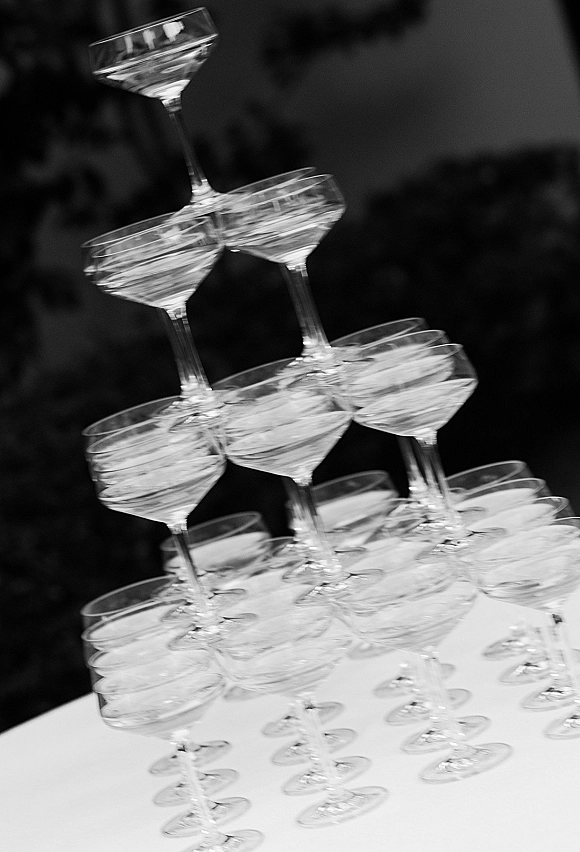 Champagne tower of stacked coupe glasses filled with champagne on a white table indoors, with guests softly blurred in background