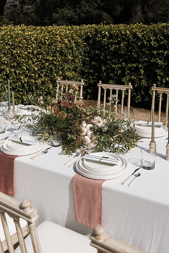 Reception tablescape with a blush table runner, garlic centerpiece and tapered candles on white linen, set outdoors beside lush garden hedges