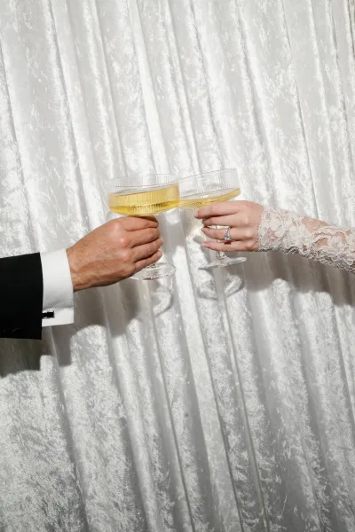 Champagne toast with champagne coupe glasses as a couple clinks, her engagement ring catching the light while bubbles rise in the coupes.