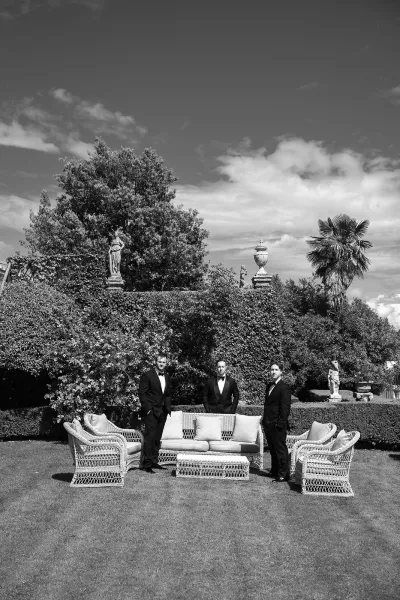 Groomsmen portrait with the party gathered around lounge furniture in a garden framed by tall hedges, trees, and classical statues.
