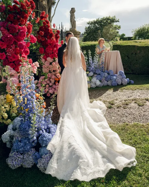 Wedding vows at a garden wedding ceremony beneath a floral arch, the couple stand together on a gravel path near hedges and a wrought iron gate.