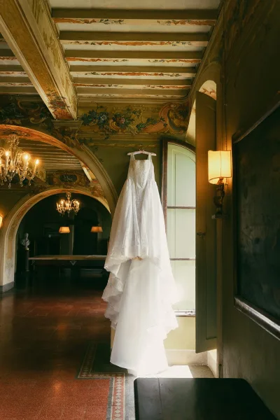 Wedding dress on a hanger in a vintage interior with a painted ceiling, chandeliers, an arched doorway, and light spilling from an open window.