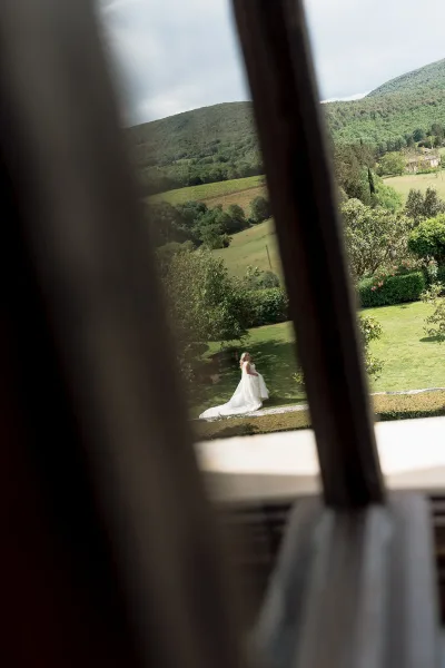Bridal portrait of a bride walking in garden along the garden path, lifting a long dress train, with trees and rolling hills under a cloudy sky.
