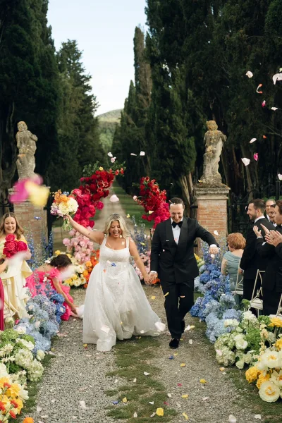 Ceremony recessional with a petal toss exit as the couple walks, guests showering them with flower petals amid garden trees and brick pillars.