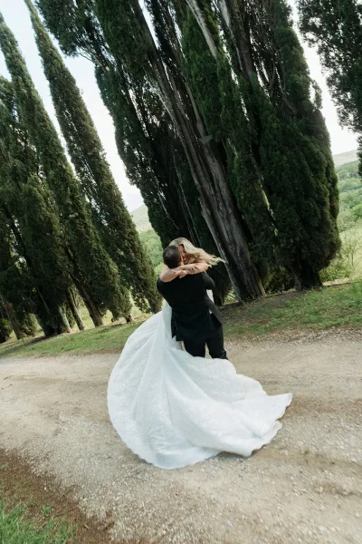 Couple portrait as the bride and groom embrace on a gravel path lined with cypress trees, her classic white gown trailing, hills beyond.